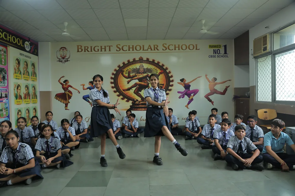 A student performing a classical dance step in front of the Nataraja mural while her class watches